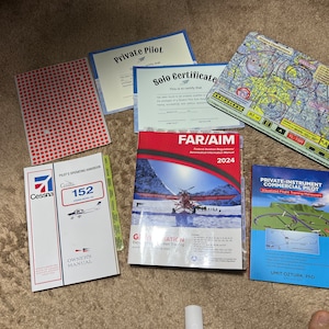 May include: A collection of aviation training materials including a Cessna 152 Pilot's Operating Handbook, a FAR/AIM Federal Aviation Regulations Aeronautical Information Manual, a Private Instrument Commercial Pilot Flight Training Manual, a Private Pilot certificate, a Solo Certificate, and a sectional chart.