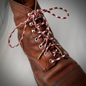 Puede incluir: Botas de trabajo de cuero marr&oacute;n con cordones rojos, blancos y verdes. Los cordones est&aacute;n atados en un patr&oacute;n de cruz.