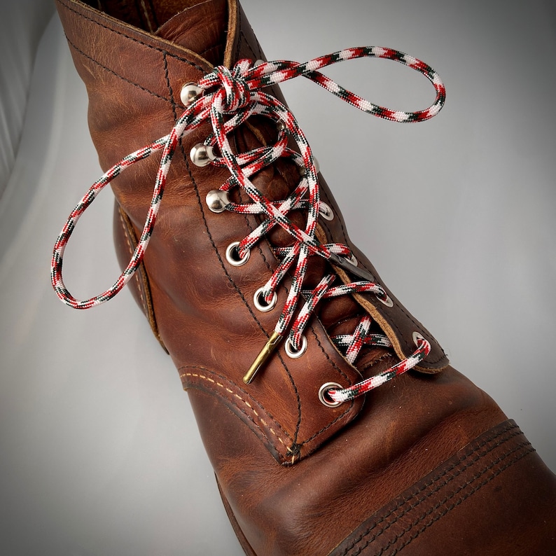 Puede incluir: Botas de trabajo de cuero marr&oacute;n con cordones rojos, blancos y verdes. Los cordones est&aacute;n atados en un patr&oacute;n de cruz.