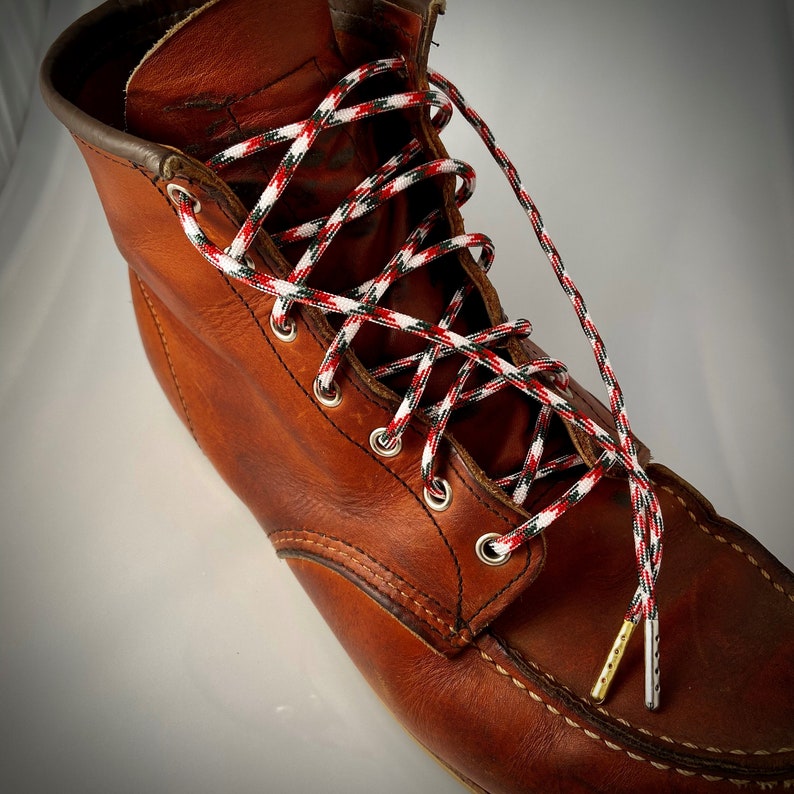 Puede incluir: Botas de trabajo de cuero marr&oacute;n con cordones rojos, blancos y verdes. Los cordones est&aacute;n atados en un patr&oacute;n de cruz.