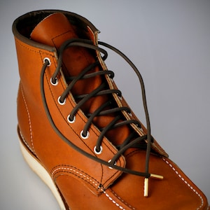 Könnte beinhalten: Nahaufnahme eines braunen Lederstiefels mit dunkelgrünen Schnürsenkeln. Der Stiefel hat eine weiße Sohle, weiße Nähte und Metallösen. Die Schnürsenkel sind kreuzweise gebunden.