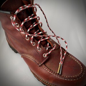 Puede incluir: Botas de trabajo de cuero marr&oacute;n con cordones de patr&oacute;n rojo, blanco y negro. Los cordones est&aacute;n atados en un patr&oacute;n de cruz y se extienden m&aacute;s all&aacute; de la parte superior de la bota.