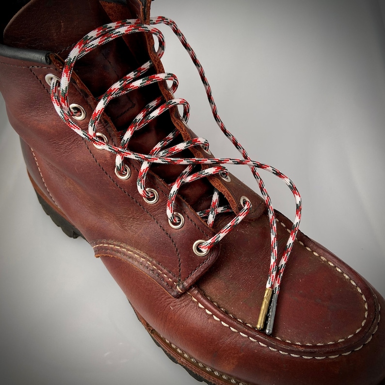 Puede incluir: Botas de trabajo de cuero marr&oacute;n con cordones de patr&oacute;n rojo, blanco y negro. Los cordones est&aacute;n atados en un patr&oacute;n de cruz y se extienden m&aacute;s all&aacute; de la parte superior de la bota.