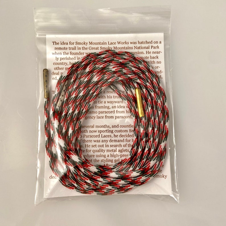 Puede incluir: Primer plano de una cuerda de paracord roja, blanca y negra con puntas de metal dorado. La cuerda est&aacute; enrollada y anudada. El texto "The idea for Smoky Mountain Lace Works was hatched on a remote trail in the Great Smoky Mountains National Park when the founder was on a solo excursion. He nearly perished in the wilderness, but he made it back with no injuries. He had a rescue rope with his trusty paracord to tie a wayward branch to his framing, an idea was born. Smoky Mountain Lace Works is a small business that makes custom paracord laces from paracord.  " es visible en el fondo.