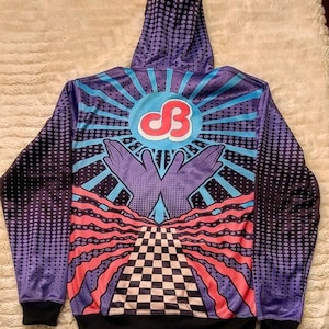 Puede incluir: Una sudadera con capucha de color morado y azul con un diseño gráfico que presenta una pirámide a cuadros, dos manos levantadas y un sol con las letras "DB" en un círculo.