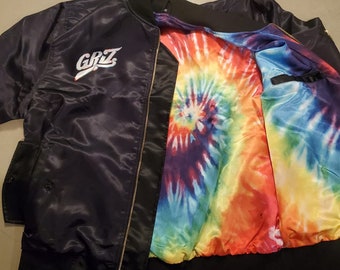 Könnte beinhalten: Eine schwarze Satin-Bomberjacke mit einem Batik-Futter. Die Jacke hat einen Reißverschluss und ein weiß gesticktes Logo mit der Aufschrift "GRZ".