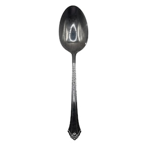 Puede incluir: Una cuchara plateada con un mango largo y un cuenco ovalado. El mango tiene un diseño decorativo cerca de la base y está acanalado a lo largo. La cuchara está sobre un fondo blanco.