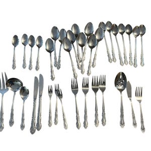 May include: A collection of ornate silver-colored flatware, including forks, spoons, knives, and serving utensils. The silverware features detailed floral patterns on the handles. The arrangement includes various sizes of spoons and forks, suitable for a formal dining setting.