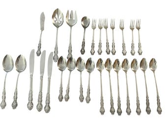 May include: A collection of ornate silver-colored flatware, including spoons, forks, knives, and serving utensils. The handles feature a detailed floral design. The set is arranged on a white surface, showcasing the intricate patterns.