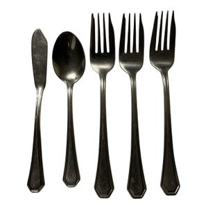 May include: A collection of five stainless steel cutlery items: a butter knife, a spoon, and three forks. The utensils have a simple, elegant design with a polished finish. The handles have a geometric shape at the base.