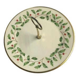May include: A two-tiered serving tray with a festive holly and berry design. The white ceramic tray features a gold rim and a silver handle. Green holly leaves and red berries encircle the edge, ideal for festive occasions.