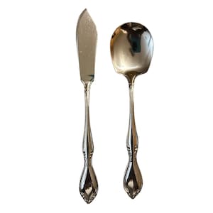 May include: A butter knife and a spoon with ornate handles. The knife has a wide, rounded blade. The spoon has a rounded bowl. Both utensils are made of a shiny, silver-colored metal. The handles have a decorative, curved design.