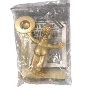 May include: A gold-colored Homer Simpson figure, holding a large, round object, packaged in a clear plastic bag. The figure is smiling with arms outstretched. The packaging includes a printed advertisement with the text "Berry Squirts".