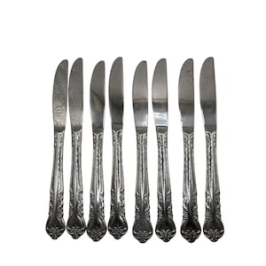 May include: A set of eight silver butter knives. The knives have a curved blade and ornate floral detailing on the handles. The blades are smooth and shiny, reflecting the light. The knives are arranged in a row.