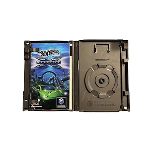 Puede incluir: Caja de juego Nintendo GameCube negra abierta con la carátula del juego "Hot Wheels Velocity X" visible. La carátula presenta un coche de carreras verde, el logotipo del juego y un fondo de ciudad. La caja está abierta, revelando el soporte del disco.
