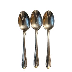 May include: Three silver spoons with elongated oval bowls and ornate, ridged handles. The spoons are arranged vertically against a white background, showcasing their polished surfaces and elegant design. The handles have a decorative gold-colored accent at the base.