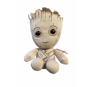 May include: A plush toy of the character Groot from Guardians of the Galaxy. The doll is beige with a textured appearance, large black eyes, and a small smile. It has a detailed head and arms, and is sitting.