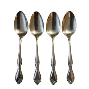 May include: Four silver spoons with an oval bowl and ornate handle design. The spoons are arranged in a row against a white background. The spoons are made of a shiny, reflective metal.