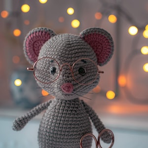 May include: A gray crocheted mouse wearing pink ears and round glasses, holding a pair of gold scissors. The mouse is standing on a wooden base.