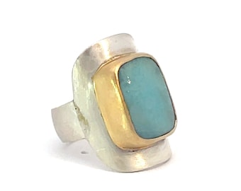 May include: A silver ring with a gold setting and a light blue gemstone. The ring has a unique, curved design.