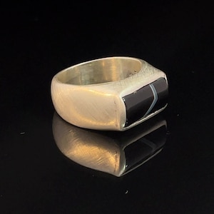 Könnte beinhalten: Ein silberner Ring mit einem schwarzen und weißen Steineinlege. Der Ring hat ein schlichtes Design mit einer flachen Oberseite und abgerundeten Seiten.