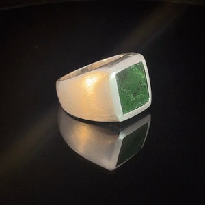 Könnte beinhalten: Ein silberner Ring mit einem quadratischen, grünen Stein. Der Ring hat eine gebürstete Metalloberfläche und eine polierte, reflektierende Oberfläche. Der grüne Stein ist in einer quadratischen Lünette gefasst.