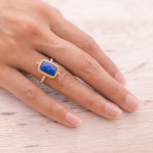 Könnte beinhalten: Ein goldener Ring mit einem blauen Edelstein, der in einer quadratischen Form gefasst ist. Der Ring befindet sich an einem Finger einer Hand, die auf einer hellen Holzoberfläche liegt.