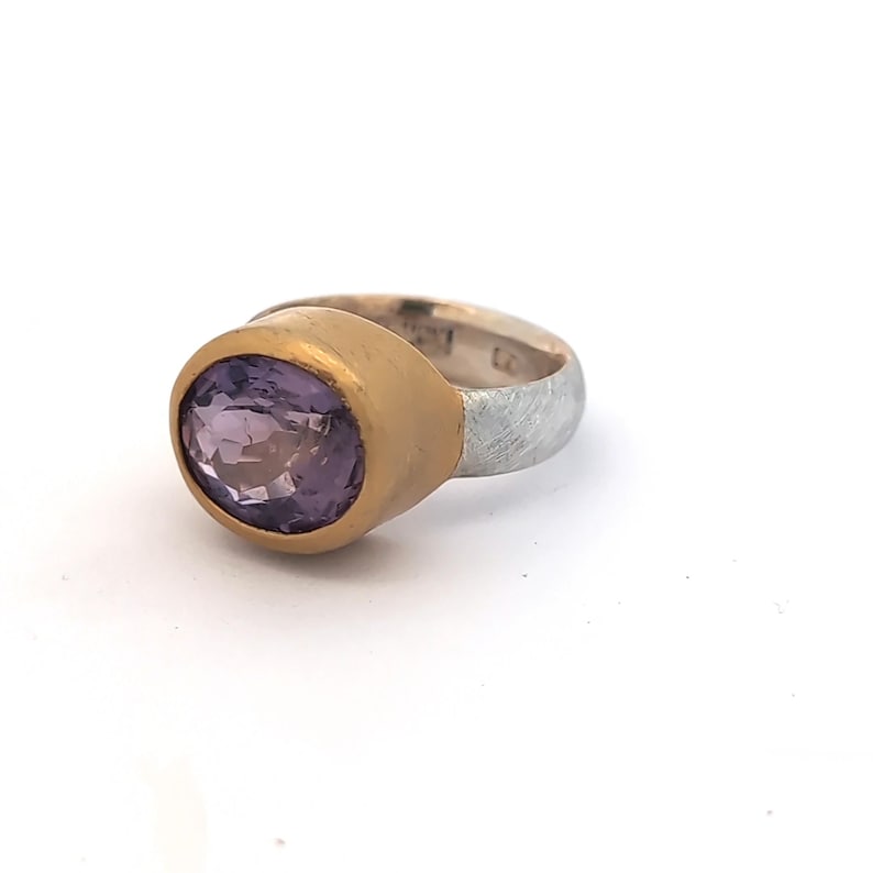 May include: An oval amethyst gemstone ring. The ring features a gold bezel and a silver band. The amethyst is a light purple hue, and the gold bezel has a brushed finish. The silver band has a textured appearance. A piece of jewellery.