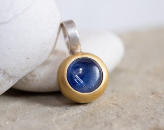 May include: A gold and silver pendant with a round blue gemstone. The pendant is set on a white background.