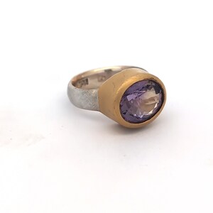 May include: A silver ring with a gold bezel setting holding a large, oval-cut, purple amethyst gemstone. The ring has a textured silver band and a gold-coloured bezel that surrounds the gemstone.