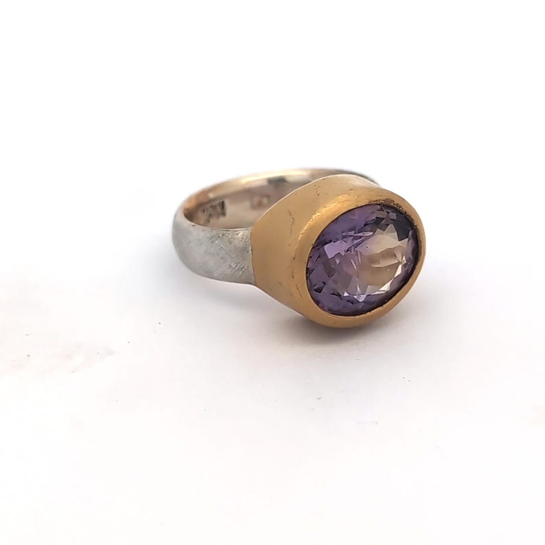 May include: A silver ring with a gold bezel setting holding a large, oval-cut, purple amethyst gemstone. The ring has a textured silver band and a gold-coloured bezel that surrounds the gemstone.