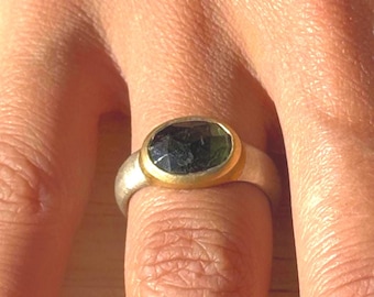 May include: A silver ring with a dark green oval gemstone set in a gold bezel. The ring is on a finger, showcasing the contrast between the silver band and the gold setting.