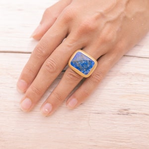 Könnte beinhalten: Ein goldfarbener Ring mit einem rechteckigen, tiefblauen Stein. Der Ring liegt auf einer hellen Holzoberfläche. Der Stein hat eine strukturierte Oberfläche, und das Design des Rings ist schlicht und elegant.