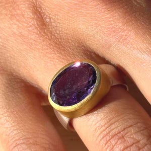 May include: A gold-plated ring with a large, oval-cut, deep purple gemstone. The ring's band is silver, and the gemstone is set in a gold bezel. The ring is displayed on a finger.