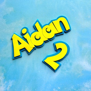 Op de afbeelding: Gele schuimletters die "Aidan" spellen met een gele schuimcijfer "2" op een blauwe achtergrond.