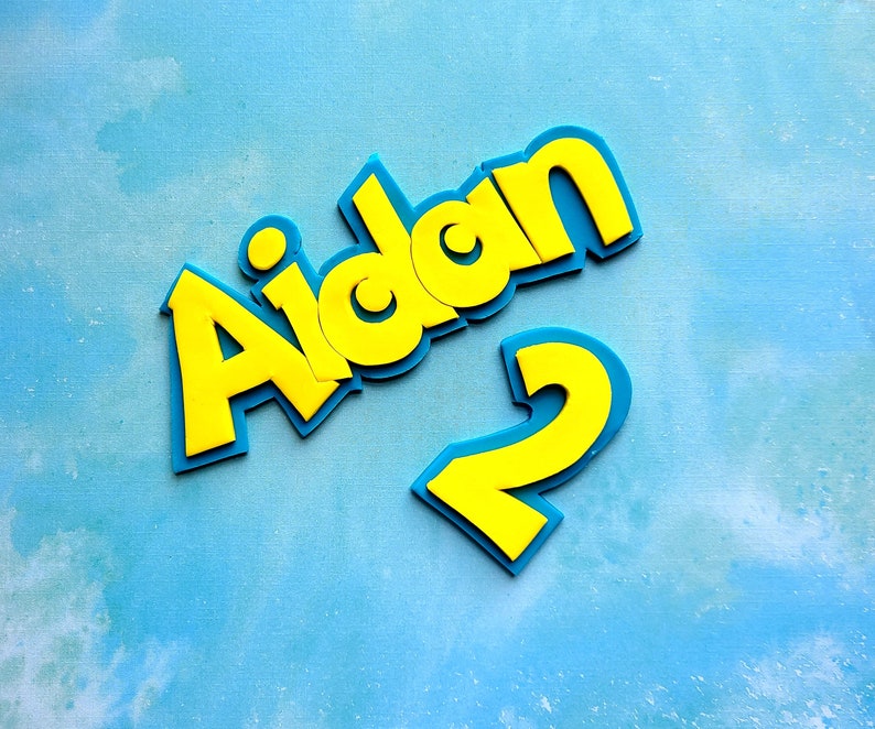 Op de afbeelding: Gele schuimletters die "Aidan" spellen met een gele schuimcijfer "2" op een blauwe achtergrond.