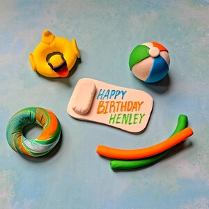 Op de afbeelding: Een set van vijf fondant caketoppers met een zomerthema. De toppers omvatten een strandhanddoek, een strandbal, een zwemband, een reddingsboei en een paar zwembadnoedels. De strandhanddoek heeft de tekst "Happy Birthday Henley" erop.