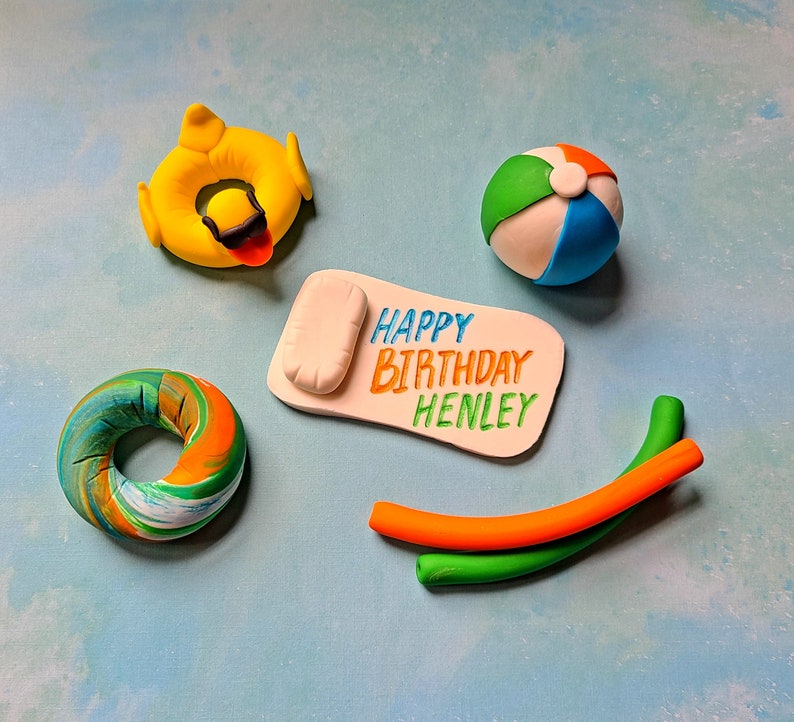 Op de afbeelding: Een set van vijf fondant caketoppers met een zomerthema. De toppers omvatten een strandhanddoek, een strandbal, een zwemband, een reddingsboei en een paar zwembadnoedels. De strandhanddoek heeft de tekst "Happy Birthday Henley" erop.