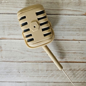 May include: A beige vintage-style microphone with a black grill and a long, thin, beige stem. The microphone is sitting on a light wood surface.
