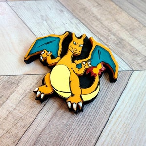 Op de afbeelding: Een houten figuur van een cartoon-draak met oranje en gele schubben, blauwe vleugels en een zwarte omtrek. De draak staat met gespreide vleugels.
