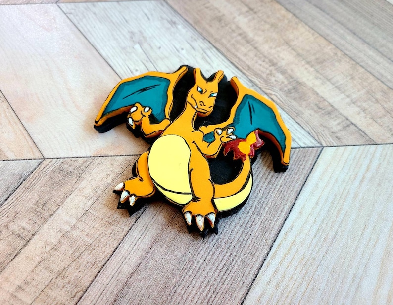 Op de afbeelding: Een houten figuur van een cartoon-draak met oranje en gele schubben, blauwe vleugels en een zwarte omtrek. De draak staat met gespreide vleugels.