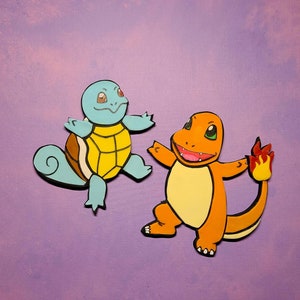 Op de afbeelding: Een blauwe en gele cartoon schildpad en een oranje cartoon hagedis met een vlammende staart. De schildpad heeft een schild met een geel midden en een blauw lichaam. De hagedis heeft een geel lichaam en een rode vlammende staart.