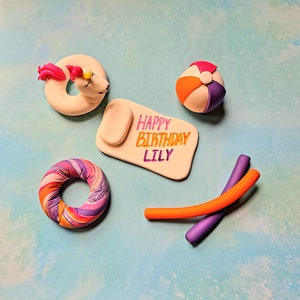 Op de afbeelding: Een set caketoppers voor een verjaardagsfeest met een zomerthema. De toppers omvatten een eenhoorn die op een opblaasbaar ringetje rijdt, een strandbal, een zwembadnoodle, een kleurrijk opblaasbaar ringetje en een wit bord met de tekst "Happy Birthday Lily".