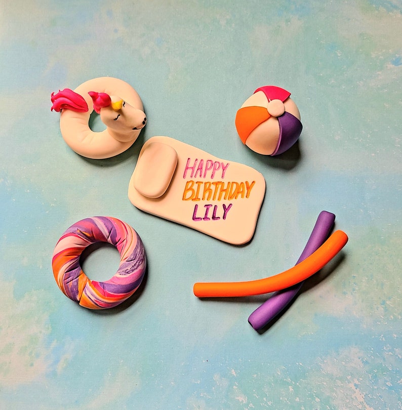 Op de afbeelding: Een set caketoppers voor een verjaardagsfeest met een zomerthema. De toppers omvatten een eenhoorn die op een opblaasbaar ringetje rijdt, een strandbal, een zwembadnoodle, een kleurrijk opblaasbaar ringetje en een wit bord met de tekst "Happy Birthday Lily".