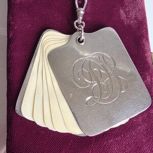 May include: A silver-toned metal dance card or notepad featuring a series of cream-coloured, thin, rectangular pages fanned out. The front cover is engraved with an ornate, cursive monogram of the letters DJ and includes a small metal swivel clasp.