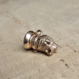 May include: A gold-colored dog pendant with a detailed head and collar. The dog's eye is a small red stone. The pendant is likely for a necklace or bracelet, and it is a vintage or antique piece.