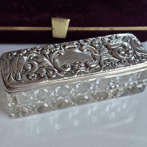 May include: A rectangular glass box with a diamond-cut pattern on the sides and a hinged silver lid. The lid features an intricate embossed design of scrolling foliage and two birds flanking a central smooth, unengraved rectangular cartouche.