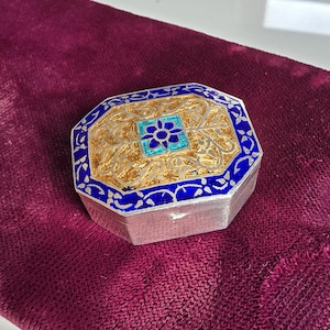 May include: An octagonal silver-toned box featuring a lid with a central blue and turquoise floral motif surrounded by intricate gold-coloured filigree patterns and a deep blue enamel border. The sides of the box are plain silver-toned metal.