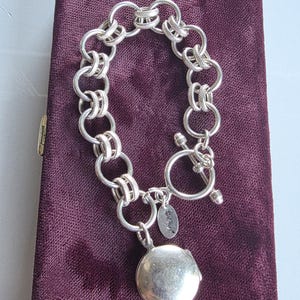 May include: A silver-toned chain bracelet featuring alternating smooth and textured circular links. The bracelet includes a toggle clasp and a round, smooth silver-toned locket charm hanging from a small jump ring, accompanied by a small oval tag.