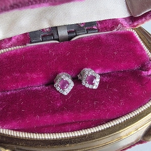 May include: A pair of stud earrings featuring a square-cut pink gemstone at the centre, surrounded by a halo of small clear stones set in a silver-toned metal. The earrings are displayed inside a vintage-style jewellery box with magenta velvet lining.